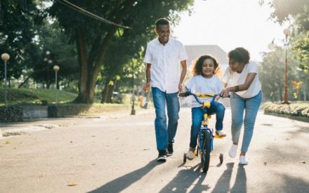 A mother and father help their child ride a bicycle outside