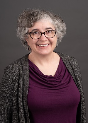 A smiling white woman with grey hair and glasses in a purple top and dark grey sweater against a grey background