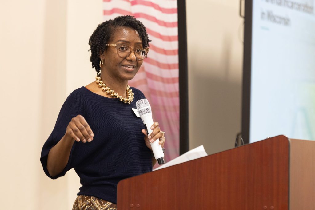 A woman holding a microphone and wearing glasses gestures at a podium
